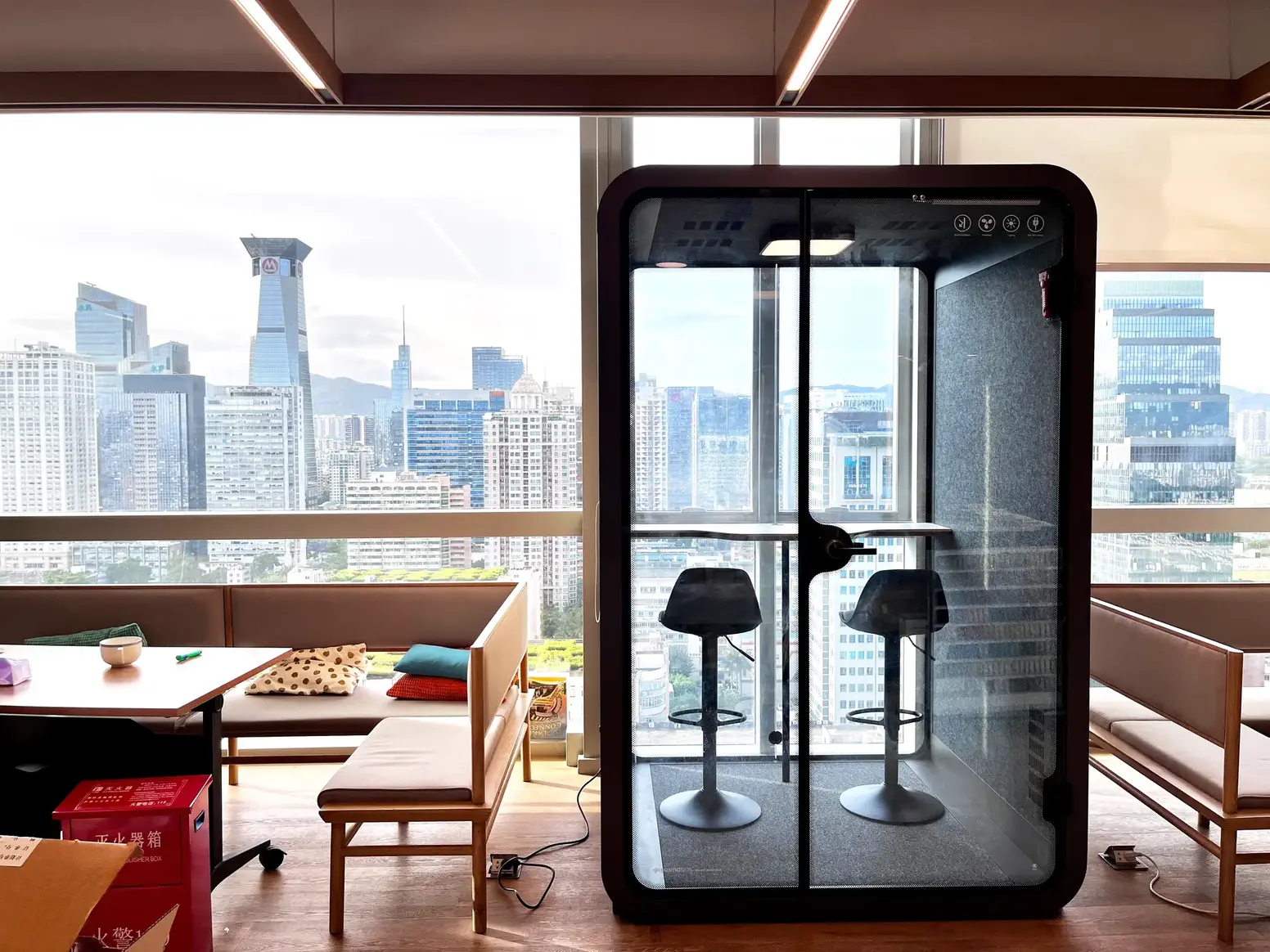 City-view office lounge with a black Soundbox SR two-person pod, two stools and a shared desk.